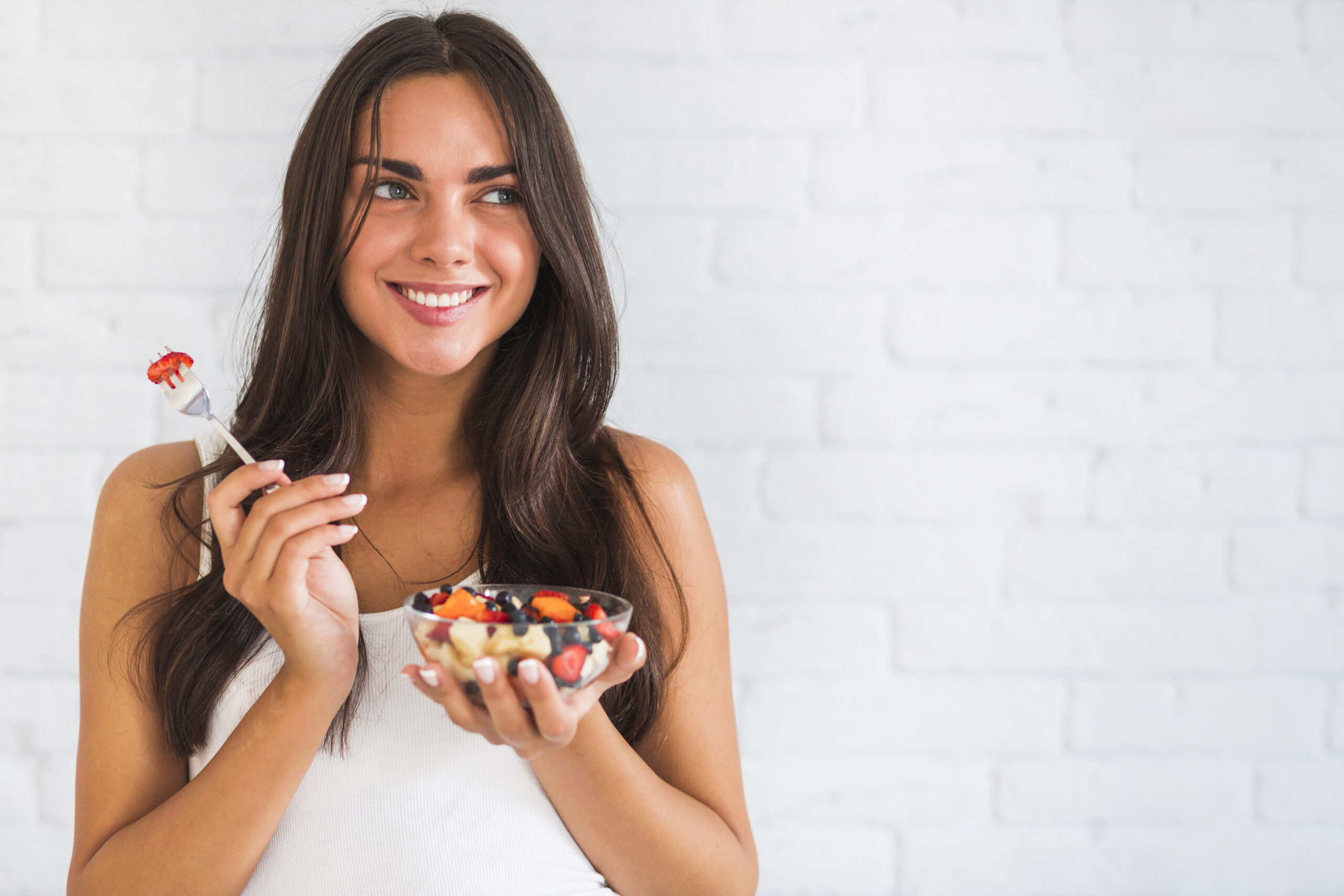 thoughtful-smiling-young-woman-holding-bowl-of-fruit-salad